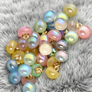 Pode incluir: Uma coleção de botões iridescentes em várias cores, incluindo rosa, amarelo, verde e azul. Cada botão tem uma argola dourada para fixar ao tecido. Os botões são redondos e têm um acabamento brilhante e perolado.