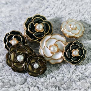 May include: A collection of flower-shaped decorative buttons. The buttons feature a pearl center and are in various colors, including black, white, and bronze, with gold-colored trim. The buttons are arranged on a gray, textured surface.