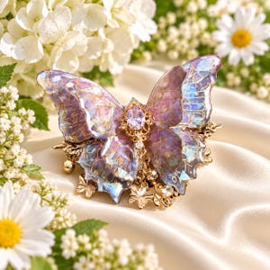 May include: A butterfly hair clip featuring translucent purple and blue wings with gold-toned vein detailing. The center is adorned with a faceted oval lavender gemstone and small dangling gold butterfly charms and clear crystals on a metal base.