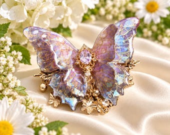 May include: A butterfly hair clip featuring translucent purple and blue wings with gold-toned vein detailing. The center is adorned with a faceted oval lavender gemstone and small dangling gold butterfly charms and clear crystals on a metal base.