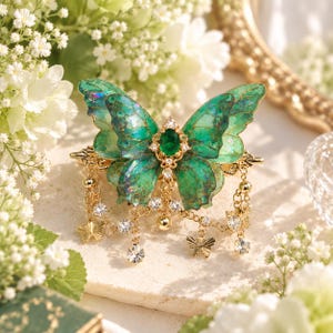 May include: A hair clip featuring a large, iridescent green butterfly with translucent wings. The center is adorned with an oval emerald-colored stone surrounded by clear crystals. Gold-toned chains hang from the base, accented with small metal butterfly charms and clear crystal beads.