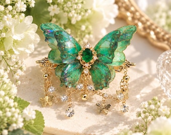 May include: A hair clip featuring a large, iridescent green butterfly with translucent wings. The center is adorned with an oval emerald-colored stone surrounded by clear crystals. Gold-toned chains hang from the base, accented with small metal butterfly charms and clear crystal beads.