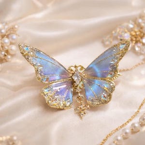 May include: A butterfly-shaped brooch featuring translucent blue and purple wings with gold glitter edges and embedded clear crystals. A central marquise-cut crystal sits at the body, with a small dangling crystal charm hanging from the bottom center.