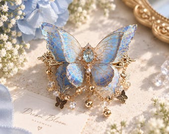May include: A gold-toned hair clip featuring a large, translucent blue butterfly with intricate gold vein detailing. The center is adorned with a clear oval gemstone, and the base includes dangling gold chains with small butterfly charms and clear beads.