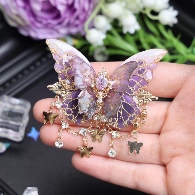 May include: A butterfly-shaped brooch featuring translucent purple and white wings with gold-toned filigree detailing. The center is adorned with a faceted light purple gemstone, and the base features dangling gold-toned butterfly charms and clear crystals.
