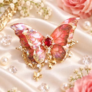 May include: A decorative hair clip featuring a large, translucent butterfly with pink and gold iridescent wings. The center of the butterfly is adorned with an oval red gemstone surrounded by small clear crystals, with a small gold butterfly charm.