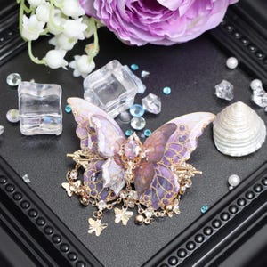 May include: A butterfly-shaped hair accessory featuring translucent purple and gold-toned wings with intricate vein patterns. The center is adorned with a faceted pink oval stone and clear crystals, with dangling gold-toned butterfly charms.