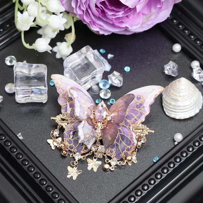 May include: A butterfly-shaped hair accessory featuring translucent purple and gold-toned wings with intricate vein patterns. The center is adorned with a faceted pink oval stone and clear crystals, with dangling gold-toned butterfly charms.