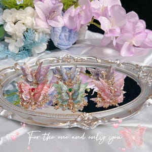 May include: Five butterfly-shaped hair clips arranged on an oval silver mirror. Each clip features translucent wings in shades of pink, blue, green, and purple, accented with small clear crystals and a central gemstone. The text reads: For the one and only you.