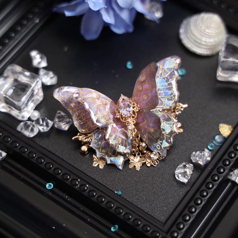 May include: A butterfly hair clip featuring translucent purple wings with intricate gold filigree patterns. The center is adorned with an oval lavender gemstone set in gold metal, with small dangling gold butterfly charms and clear crystals attached.