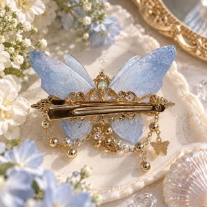 May include: A hair clip featuring a light blue butterfly with translucent, textured wings. The center is adorned with ornate gold-tone metal filigree, small clear crystals, and dangling gold-tone chains with tiny beads and a small butterfly charm.
