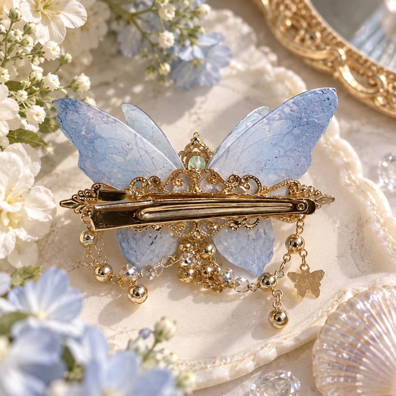May include: A hair clip featuring a light blue butterfly with translucent, textured wings. The center is adorned with ornate gold-tone metal filigree, small clear crystals, and dangling gold-tone chains with tiny beads and a small butterfly charm.