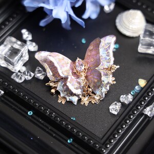May include: A butterfly hair accessory featuring translucent purple and iridescent wings with gold-toned vein detailing. The center is adorned with a faceted oval purple stone and dangling gold-toned butterfly charms and small clear beads.
