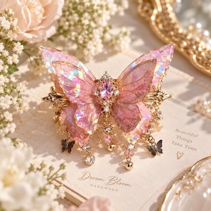 May include: A hair clip featuring a large, iridescent pink butterfly with translucent, shimmering wings. The center is adorned with a faceted pink oval gemstone and clear crystals. Small gold chains with dangling crystal beads and tiny butterfly charms hang below.