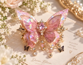 May include: A hair clip featuring a large, iridescent pink butterfly with translucent, shimmering wings. The center is adorned with a faceted pink oval gemstone and clear crystals. Small gold chains with dangling crystal beads and tiny butterfly charms hang below.