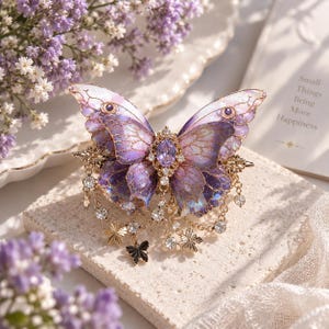 May include: A butterfly-shaped hair clip featuring translucent purple and pink wings with gold-toned veins and glitter accents. The center is adorned with an oval purple gemstone and clear crystals, with dangling gold chains and small charms.