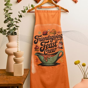 May include: Orange Friendsgiving Feast Crew apron with a pocket featuring a gravy boat illustration. The apron has the text "Friendsgiving Feast Crew" with food icons. The apron is hanging on a wooden hanger.