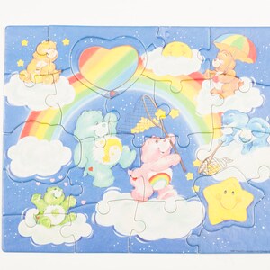 Może przedstawiać: Kolorowa układanka z Care Bears z tęczą, chmurami i gwiazdami. Care Bears bawią się siatką i balonem w kształcie serca.