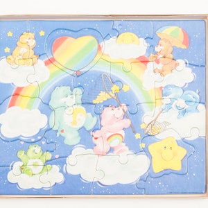 Może przedstawiać: Puzzle z Care Bears z tęczą, chmurami i gwiazdami. Puzzle przedstawia osiem Care Bears, w tym Cheer Bear, Funshine Bear i Grumpy Bear. Puzzle jest wykonane z tektury i jest odpowiednie dla dzieci w wieku od 3 lat.