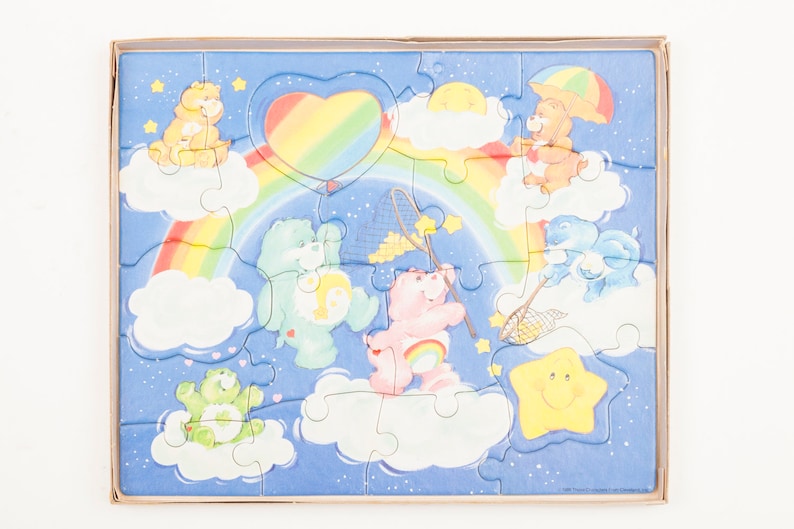 Może przedstawiać: Puzzle z Care Bears z tęczą, chmurami i gwiazdami. Puzzle przedstawia osiem Care Bears, w tym Cheer Bear, Funshine Bear i Grumpy Bear. Puzzle jest wykonane z tektury i jest odpowiednie dla dzieci w wieku od 3 lat.