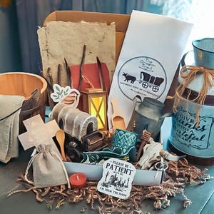 Peut inclure: Un assortiment d'objets sur le thème rustique, dont un seau en bois, une cruche à lait en métal et un petit modèle de chariot. Les articles sont disposés dans une boîte avec un fond en papier, évoquant une esthétique vintage. La boîte comprend une pancarte qui dit "Please be patient with me, I'm from the 1800s."