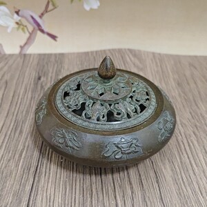 May include: A bronze-colored incense burner with a detailed, pierced lid and a small, conical finial. The body of the burner is decorated with relief patterns. The overall design suggests an Asian-inspired aesthetic.