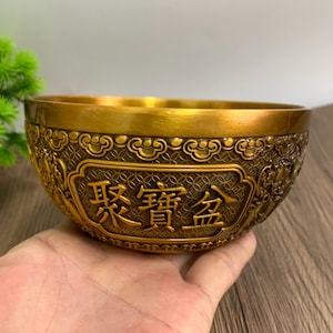 Puede incluir: Un cuenco dorado con intrincados patrones en relieve y caracteres chinos. El cuenco tiene un diseño amplio y abierto y se sostiene en una mano. El texto del cuenco está en un panel rectangular.