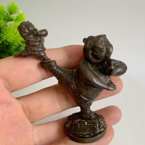 Puede incluir: Una figura de bronce de un personaje parecido a un oso en una pose juguetona, sosteniendo una figura más pequeña. La escultura tiene un diseño detallado y está montada sobre una base circular. La figura es de color marrón oscuro.