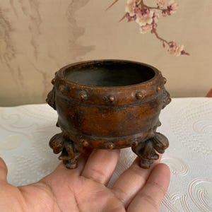 May include: A small, antique bronze-colored bowl with a rounded body and three ornate feet. The bowl has a dark interior and is decorated with small, raised studs around the rim. The feet have a detailed, floral-like design.