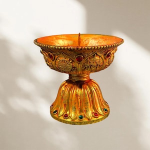 May include: A gold-coloured metal bowl with a wide, flared rim and a pedestal base. The exterior features intricate embossed patterns, including circular motifs and figures, accented with small, embedded red and green gemstones throughout.