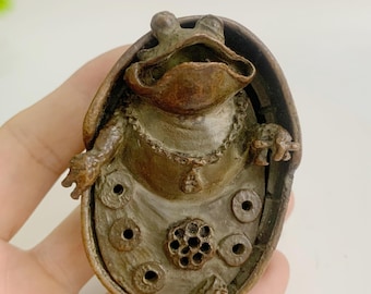 Könnte beinhalten: Bronze-Froschskulptur mit aufwendigen Details. Der Frosch sitzt in einer muschelartigen Struktur, verziert mit einer Halskette. Die Skulptur weist eine Reihe von kreisförmigen und floralen Mustern auf, die ein detailliertes Design zeigen. Die Gesamtästhetik ist antik und dekorativ.