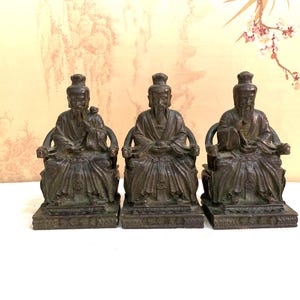 Könnte beinhalten: Drei bronzefarbene Statuen von sitzenden Figuren mit langen Bärten und traditioneller Kleidung. Jede Figur sitzt auf einem Stuhl mit Armlehnen und einem detaillierten Sockel. Die Statuen sind nebeneinander angeordnet.