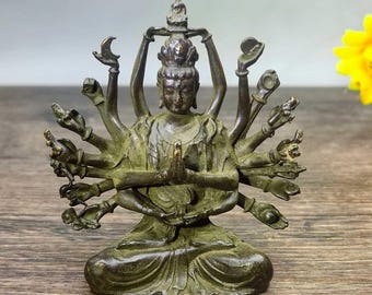 May include: Bronze-colored statue of a seated deity with multiple arms, hands clasped in prayer. The figure wears a headdress and is adorned with intricate details. The statue is on a wooden surface.
