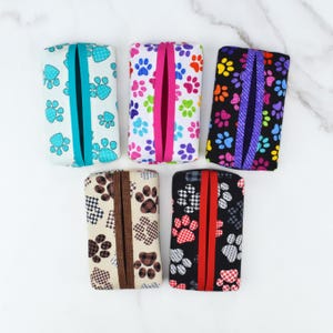 May include: Five rectangular tissue box covers with various paw print patterns. Designs include light blue, pink, orange, and black paw prints on white, black, and patterned backgrounds. Each cover has a vertical slit for tissue dispensing.