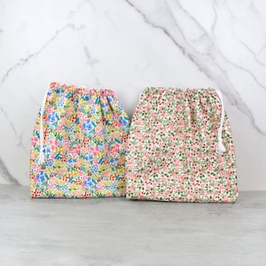 May include: Two drawstring bags with floral patterns. One bag features a colorful design with pink, blue, and yellow flowers. The other bag has a pink and green floral pattern. Both bags have white drawstrings.
