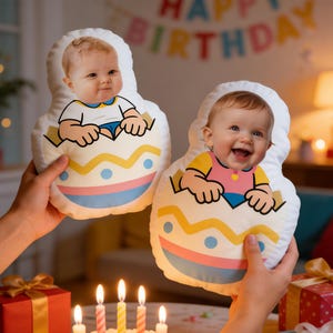 Könnte beinhalten: Zwei Plüschkissen in Eiform mit einem aufgedruckten Babygesicht. Das Baby trägt ein Hemd und scheint aus dem Ei zu schlüpfen. Die Kissen werden vor einem Geburtstagskuchen mit brennenden Kerzen gehalten.
