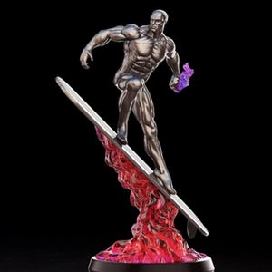 Puede incluir: Estatua plateada del Silver Surfer sobre una tabla de surf, sobre una base de energía roja y púrpura. La figura es musculosa y detallada, en una pose dinámica. La base tiene un borde negro.