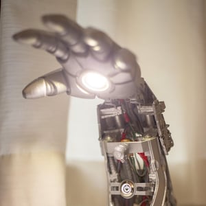Può includere: Lampada a forma di braccio robotico color argento, con una luce intensa che emana dalla mano. L'interno del braccio rivela fili e componenti meccanici. La lampada ha un pulsante di accensione e porte USB. La lampada è su una superficie bianca.