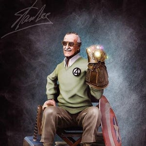 Puede incluir: Estatua de Stan Lee sentado, sosteniendo el Guantelete del Infinito. Viste un suéter verde con un emblema "4", pantalones marrones y zapatillas blancas. La base presenta una máscara de Spider-Man, un casco de Iron Man y el texto "EXCELSIOR 1922-2018."