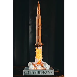 Könnte beinhalten: Ein detailliertes Modell der Saturn V-Rakete im Startzustand. Die Rakete ist hauptsächlich kupferfarben mit weißen Abschnitten und auf einem grauen Sockel montiert. Auf dem Sockel steht "SATURN V". Der Raketenausstoß wird mit orangefarbenem und weißem Licht dargestellt.