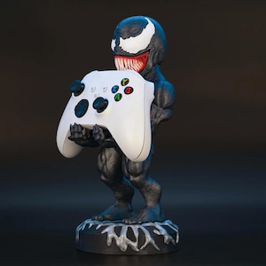 Puede incluir: Figura de Venom en blanco y negro sosteniendo un mando de Xbox blanco. La figura presenta una escultura detallada con una sonrisa amenazante y un símbolo blanco con forma de araña en la cabeza. La base es negra con un diseño blanco texturizado.