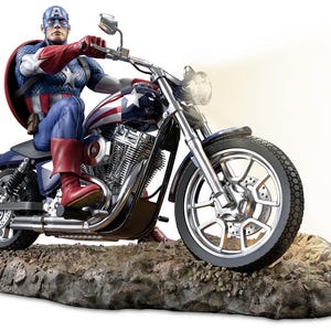 Può includere: Una statuina di Captain America su una moto. La statuina indossa un costume e un casco blu, bianco e rosso. La moto è nera e argentata con un disegno a stelle sul serbatoio. La statuina è montata su una base rocciosa.