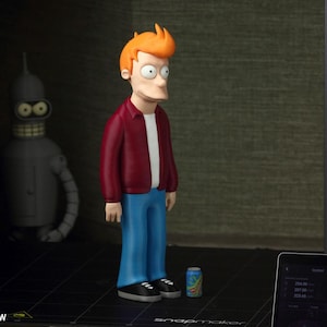 May include: A 3D-printed figurine of Philip J. Fry from Futurama, wearing a red jacket, white shirt, and blue pants. A Bender figurine is in the background. A small can and a tablet are also visible.
