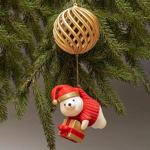 Op de afbeelding: Een gouden, bolvormige kerstversiering met een rasterontwerp hangt boven een ijsbeeroversiering. De ijsbeer draagt een rode muts en trui en houdt een cadeau vast met een rode strik. De ornamenten hangen aan een groene dennentak.