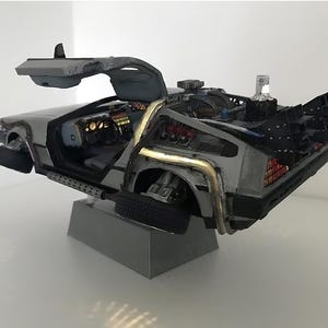 Op de afbeelding: Een gedetailleerd model van een DeLorean tijdmachine, met vleugeldeuren open. De auto is zilverkleurig met ingewikkelde details, waaronder zichtbare motoronderdelen en een futuristisch ontwerp. Het model is op een grijze basis gemonteerd.