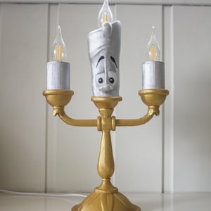 Può includere: Un candelabro dorato con tre portacandele argentati, ciascuno sormontato da una lampadina trasparente. Il portacandele centrale ha la forma di un personaggio dei cartoni animati con un volto. Un oggetto di arredamento per la casa.