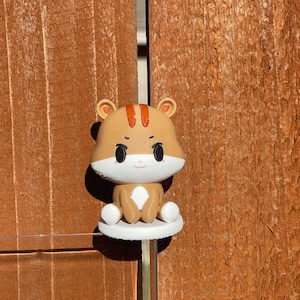 Op de afbeelding: Een charmant handgemaakt hamsterbeeldje. De hamster is bruin en wit met zwarte ogen en een glimlach. Het beeldje staat op een witte schijf, gemonteerd op een transparante staaf. De achtergrond is een houten oppervlak.