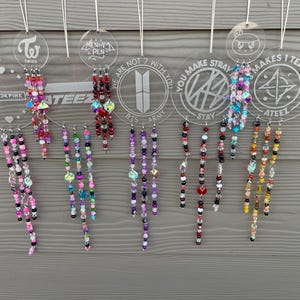 Puede incluir: Una colección de adornos de cuentas con etiquetas circulares transparentes con logotipos de bandas y texto. Los adornos tienen cuentas de varios colores, incluyendo rosa, morado, rojo y acentos iridiscentes. Las etiquetas incluyen el texto "TWICE", "BLACKPINK", "ATEEZ", "BTS ARMY" y "STRAY".