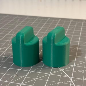 以下が含まれることがあります： 上部にハンドルが付いた2つの緑色の円筒形オブジェクト。オブジェクトは固体材料でできており、グリッドパターンを持つ灰色の表面に置かれています。オブジェクトは形状とサイズが同一です。