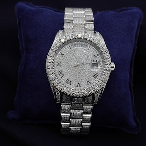 May include: A silver wristwatch with a diamond-set bezel and face. The watch has Roman numeral hour markers and a date display. The strap is also diamond-covered. The watch is displayed on a dark blue velvet surface.